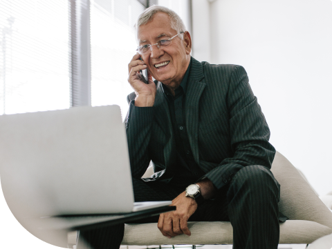 older businessman smiling while speaking on smartphone and logged into online banking portal on laptop