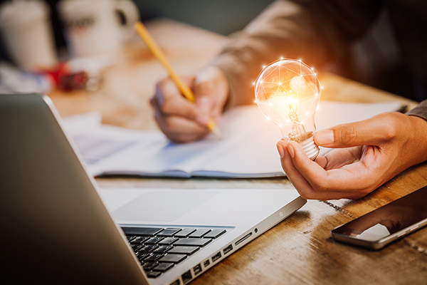 Person behind computer screen filling out paperwork and holding a lightbulb