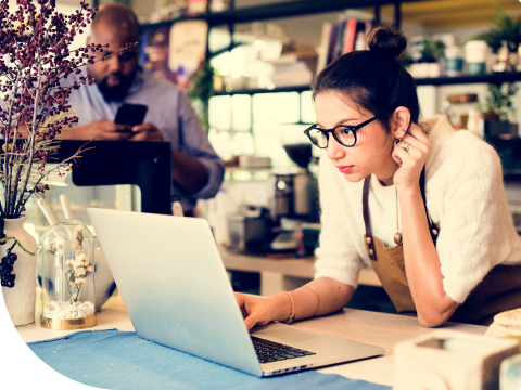 woman at desk looking at laptop with man on cellphone in background