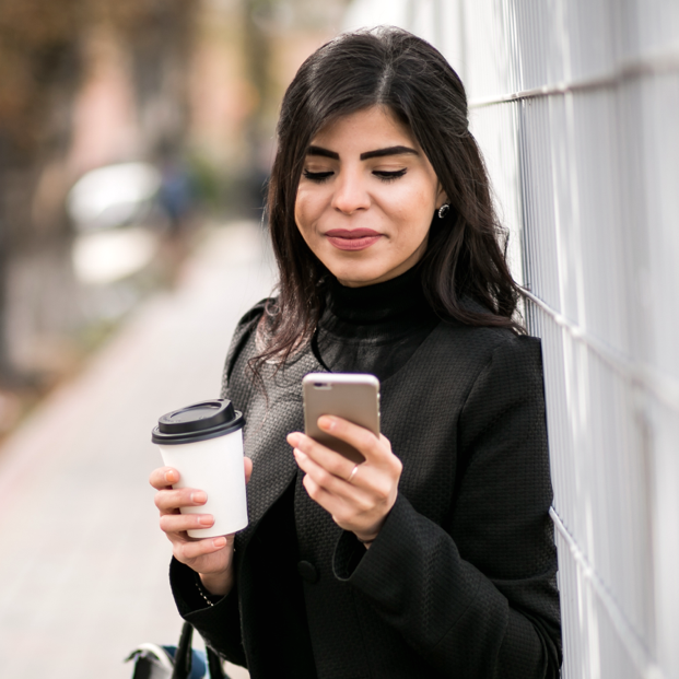 Young woman confidently looking at the BNC mobile app on her smartphone and holding a cup of coffee while outdoors.